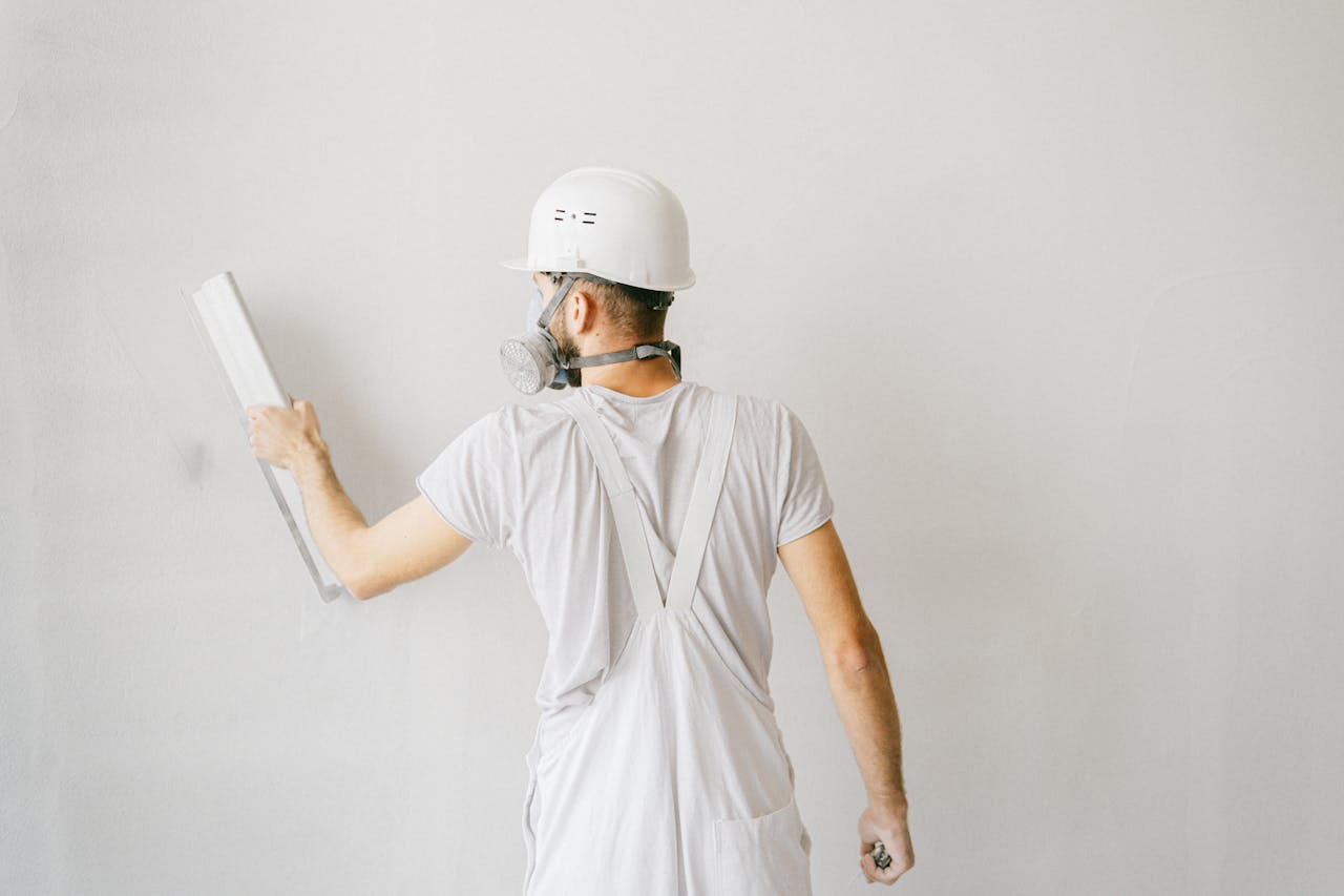 Back view of a construction worker in safety gear plastering a white wall.